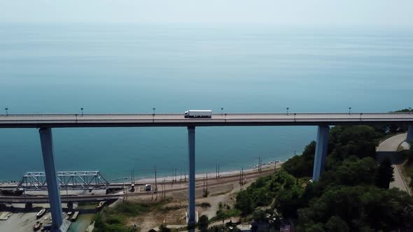 Aerial Follow Shot of White Semi Truck Moving Through High Bridge Near Blue Sea and Railway Rural