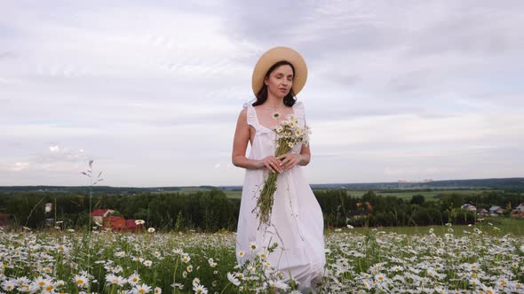 Woman in a White Dress and Hat Walks Through a Field with Daisies in the Evening at Sunset in Summer