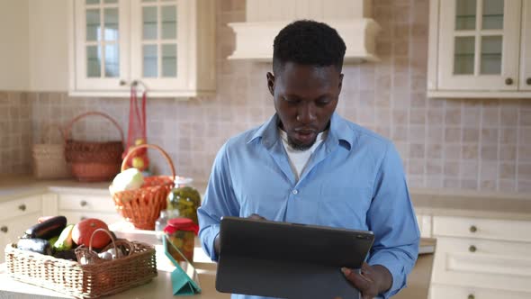 Young African American Man Surfing Internet on Tablet Standing in Kitchen with Harvest at Background