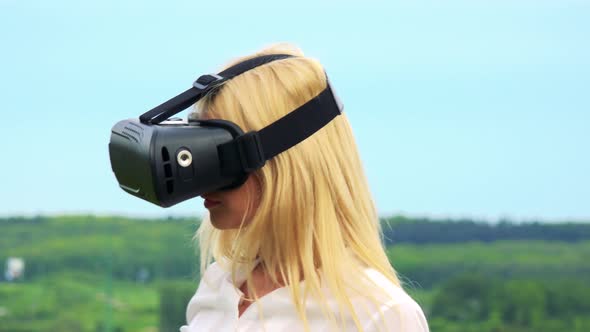 Young Attractive Blond Woman Uses Virtual Reality Glasses - Countryside and Blue Sky in Background