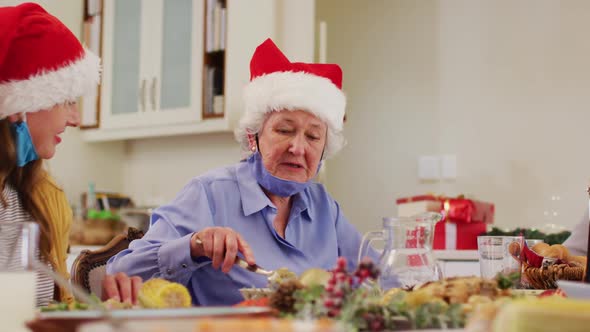 Caucasian senior woman wearing santa hat and face mask around her neck smiling while sitting on dini