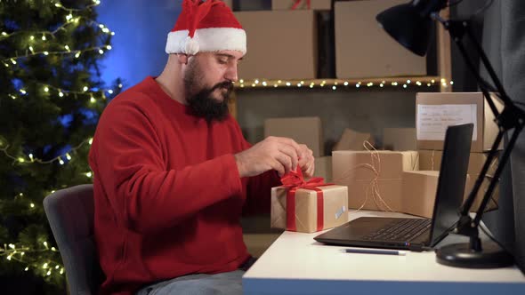 Happy Idus Bearded Santa Claus in a Suit Sits in a Home Office at a Table Among Boxes Ties a Red Bow