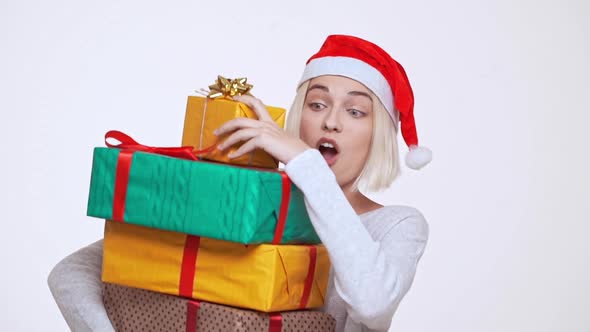 Young Pretty Blonde Girl in Christmas Hat Hardly Holding Stack of Falling Gift Boxes Over White