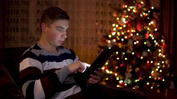 A Young Man Sits with a Tablet in Him Hands on the Background of a Christmas Tree. The Room Is