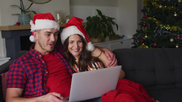 Happy caucasian couple wearing santa hats sitting on sofa and using laptop