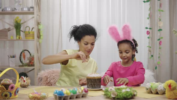 Mom and Daughter with Funny Bunny Ears Decorate Easter Cake with Sweet Candies