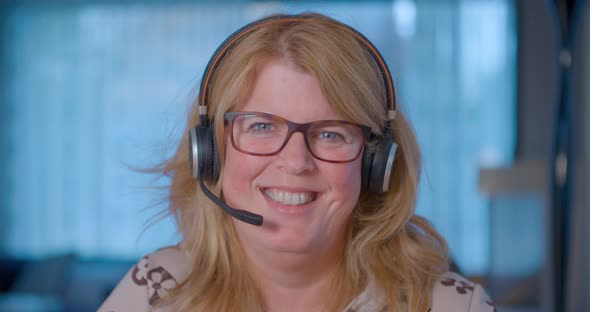 A blonde business woman is smiling at the camera while wearing a headset and glasses