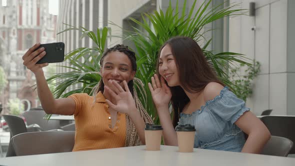 Two Happy Girlfriends Taking Photo or Have Video Chat with Friends on Their Smartphone Sitting in