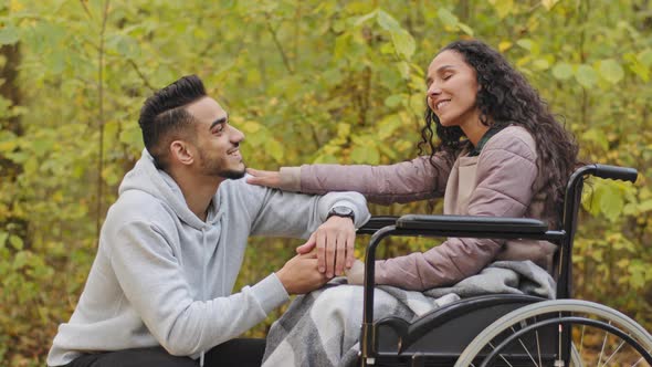 Young Hispanic Man Squatting Talking to Girl in Wheelchair Holding Hands Couple in Love Relaxing in