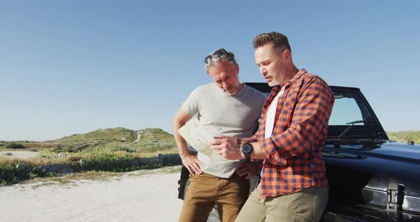 Happy caucasian gay male couple leaning on car, reading map and talking on sunny day at the beach