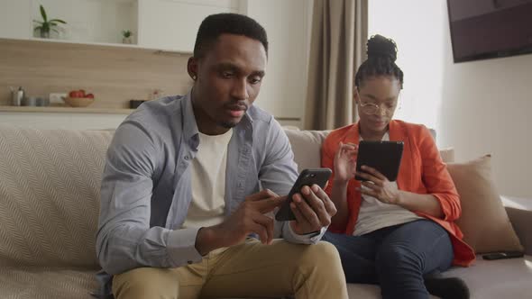 Two Young African American Adult Persons are Chilling at Home and Surfing Web on Phones and Tablets