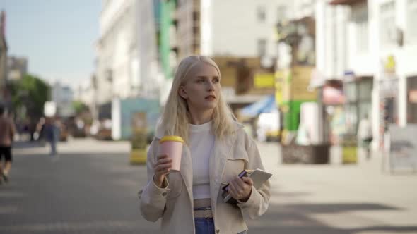 A Beautiful Young Blonde Woman Walks Along the Central Street with Books and Aromatic Coffee