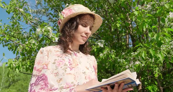 Slow Motion, Girl in Hat Sitting in Park Near a Flowering Tree Is Reading Book. Close-up, View of