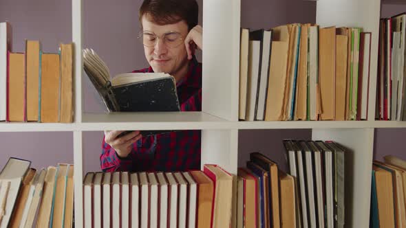 Man in Glasses Reading Book Standing Behind Bookshelf