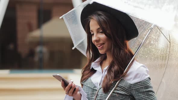 Beautiful Young Business Woman Using Smartphone on the Street in Rainy Weather, Smiling, Holding