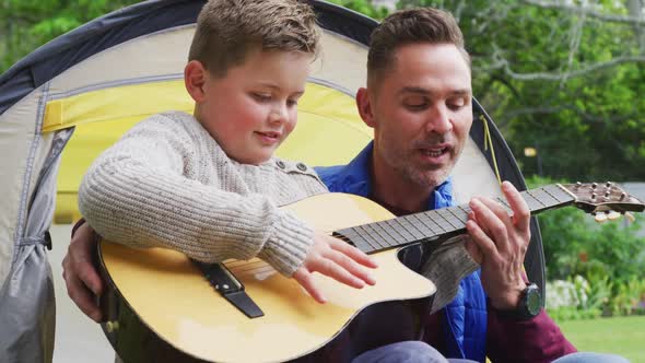 Happy caucasian father with son sitting in tent and playing guitar in garden