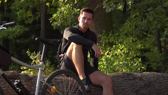 A Young Handsome Cyclist Sits on a Log Next To His Bike in a Forest and Looks Seriously