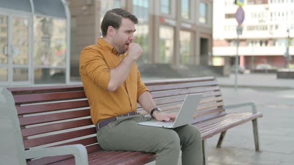 Coughing Young Man Using Laptop while Sitting Outdoor on Bench