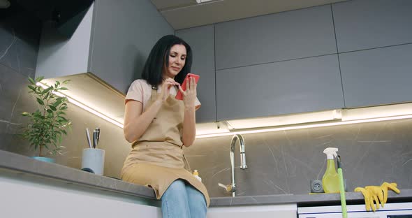 Woman in Kitchen Apron with Smartphone in Hands Chuckle while Sitting in the Kitchen at home