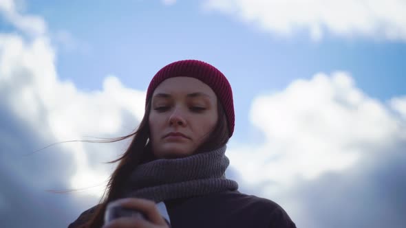 A Woman Stands on the Background of the Sky in Warm Clothes Drinks Hot Coffee