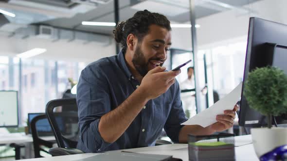 Mixed race businessman sitting at desk holding document, using smart phone and smiling