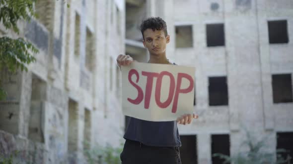 Portrait of Male Emigrant or Refugee Posing with Stop Banner at the Background of Neglected Urban