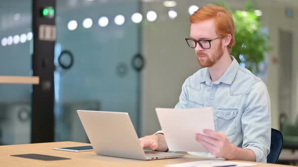 Casual Redhead Man Working on Laptop with Documents in Office 