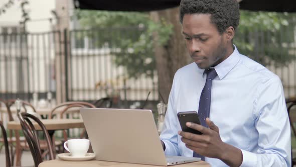 African Businessman Using Smartphone and Laptop in Outdoor Cafe