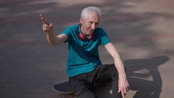 High Angle View Positive Senior Sportsman Sitting on Skateboard on Sunny Park Alley Saluting in Slow