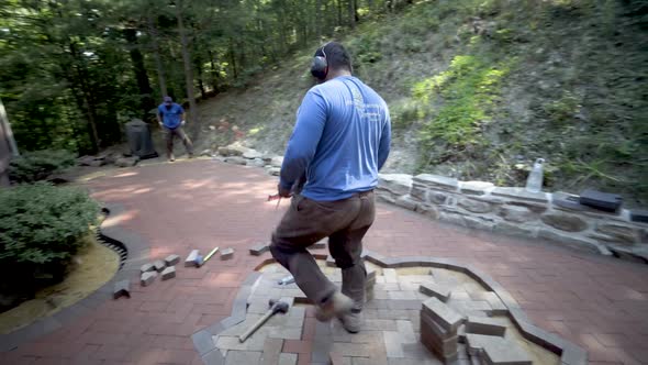 Brick paver artisan wearing ear protection marches over with freshly cut brick, puts it into positio