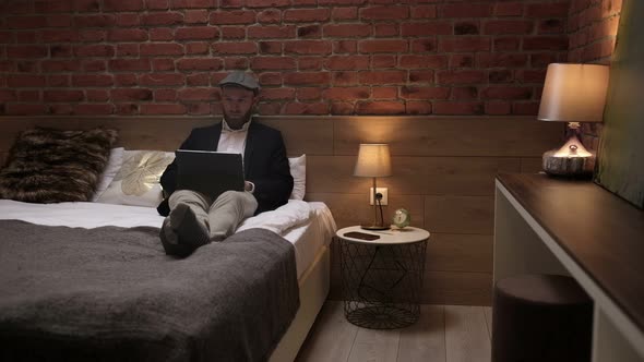 Handsome young businessman in a formal white shirt sits on a bed with a tablet in a modern room.