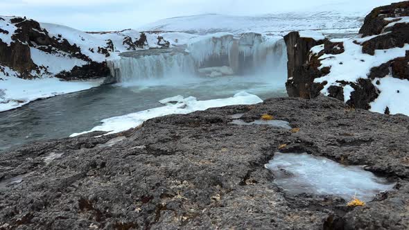 Slow motion Tilt up shot of Goðafoss Waterfall surrounded by snowy hills on Iceland during daytime