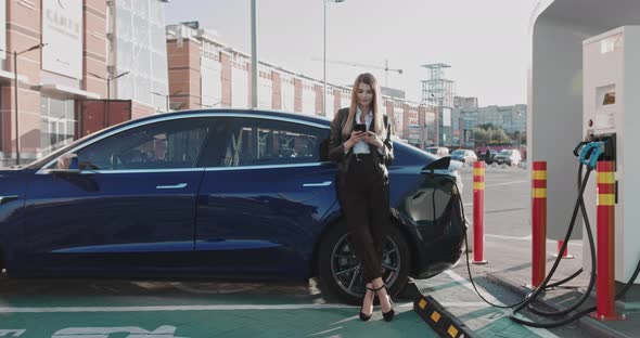 Woman Stands with Phone Near Her Electric Car and Waits When Vehicle Will