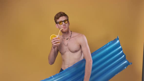 Redhaired Man in Beach Shorts with an Inflatable Mattress and a Cocktail Points His Finger to the