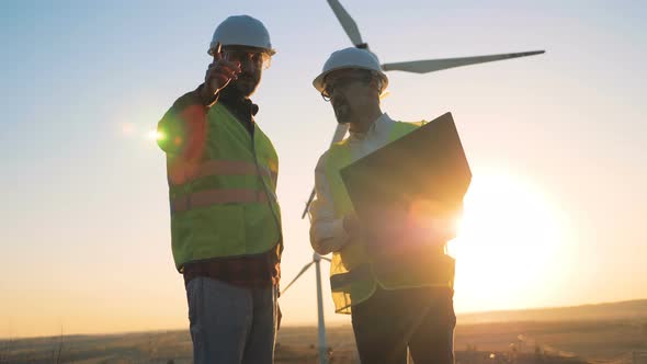 Sunset Landscape with Two Engineers Talking Beside a Set of Turbine Towers. Renewable Alternative