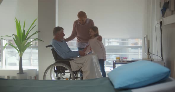 Mature Wife and Little Granddaughter Visiting Aged Disabled Man in Wheelchair at Hospital Ward