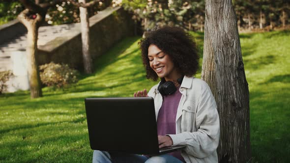 Ethnic Black Female is Smiling Waving Her Hand Chatting Online Using Laptop Sitting on Green Grass