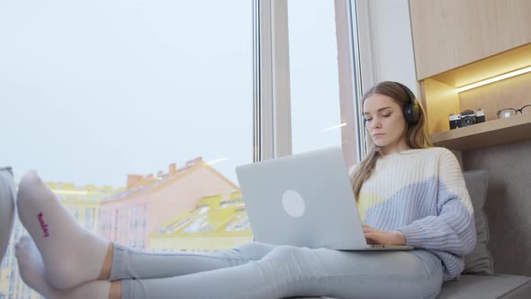 Young woman sitting by the window at home in headphones works at a laptop. Remote study. Remote work