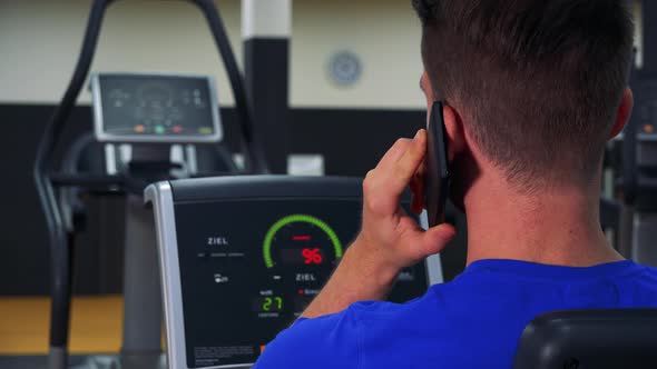 A Fit Man Trains on a Recumbent Bike in a Gym and Talks on a Smartphone - Closeup From Behind