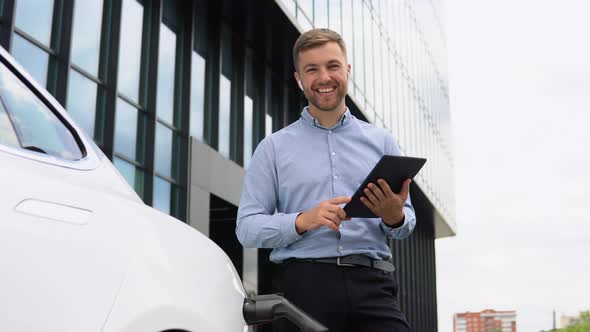 Young European Businessman with Wireless Headphones and Tablet Near Electric Car Charging the