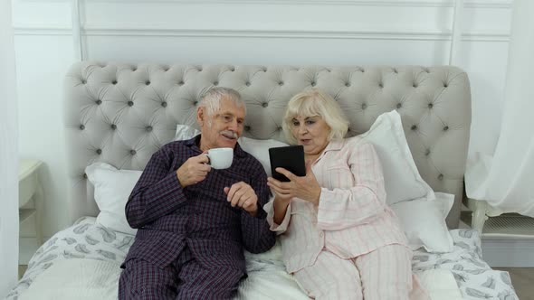 Senior Elderly Couple Wearing Pyjamas Lying on Bed Looking on Digital Tablet Laughing and Having Fun