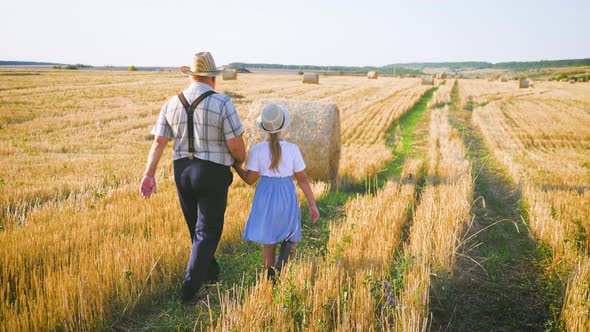 Grandfather and Granddaughter Walking Across the Field with Haystacks. Farmer Grandfather Teaches