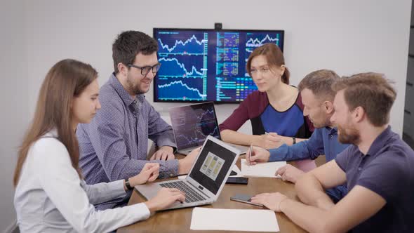 Group of Young Financial Specialists Is Sitting in Meeting Room with Display with Schedules on Wall