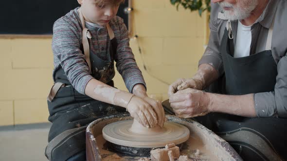 Tilt-up of Boy in Apron Shaping Clay on Throwing Wheel While Old Man Speaking Teaching
