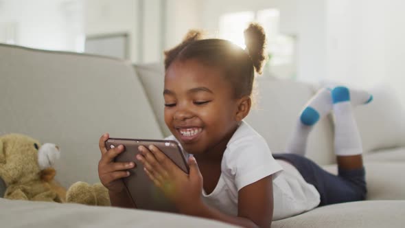 Happy african american girl lying on sofa using digital tablet at home