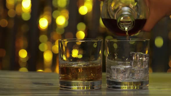 Pouring of Golden Whiskey, Cognac or Brandy From Bottle Into Glass with Ice Cubes. Shiny Background