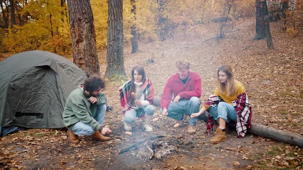 Group of Young Men and Women Smiling Talking and Roasting Sausages on Burning Campfire