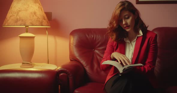 Beautiful woman with red jacket reads the book sitting on the sofa at home