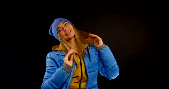 Portrait of a Blonde in a Blue Hat and a Sports Jacket on a Black Background. the Girl Smiles and