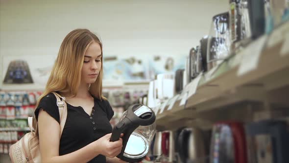 Young Pretty Woman is Inspecting Sample of Electric Kettle Near Shelf of Store